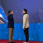 Two men stand on a stage facing each other one raises his hand in a greeting or gesture while the other listens Both wear smart casual attire against a blue tech themed backdrop
